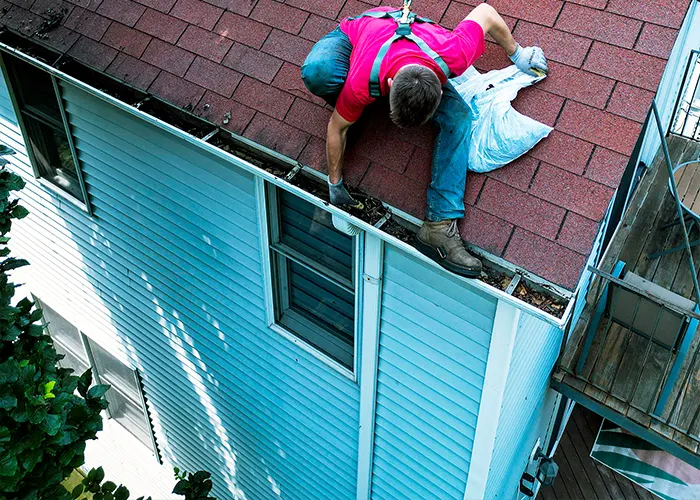 A worker in a harness clears debris from the gutter on a red shingle roof